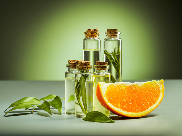 Four glass bottles with cork stoppers containing clear liquid and green leaves, positioned behind a fresh orange slice and green leaves on a light surface with a green gradient background.